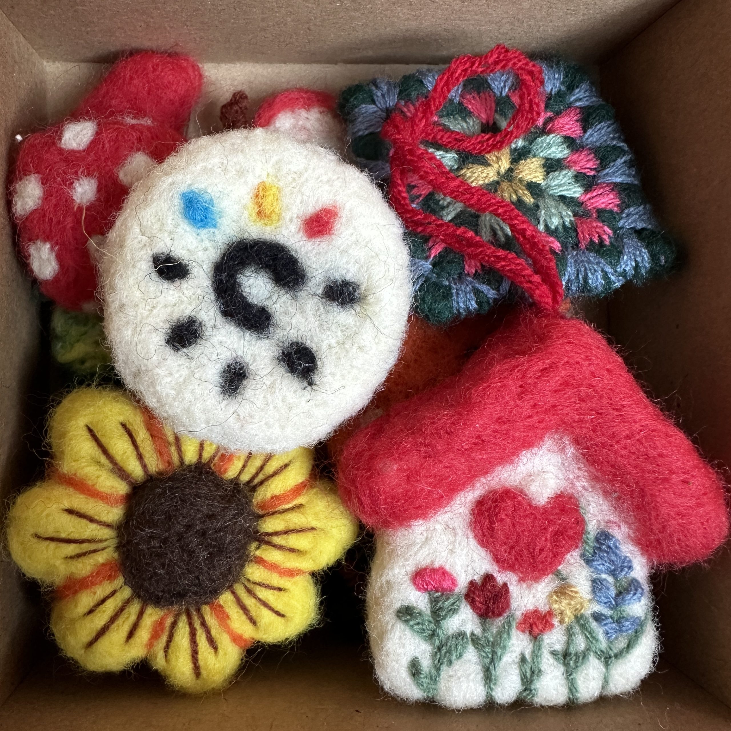 Картонена кутия, пълна с различни ръчно изработени фигурки от плъстена вълна, включително къщичка, жълт слънчоглед и абстрактни форми.