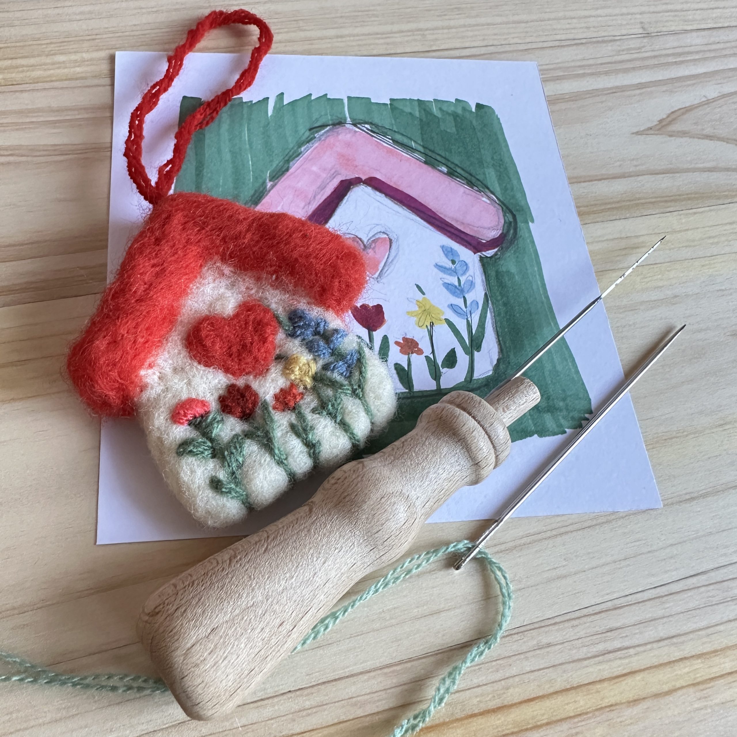 Процес на изработка чрез иглонабиване: готова мартеница от вълна, дървена игла за сухо плъстене и първоначалната цветна скица.